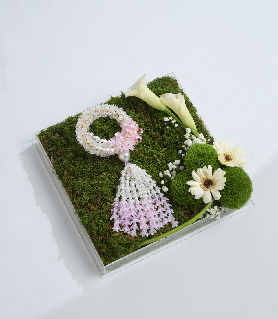 Decorative moss arrangement in square white container with white and pink flowers, calla lilies, gerberas, baby's breath, and green pompom flowers on moss base.