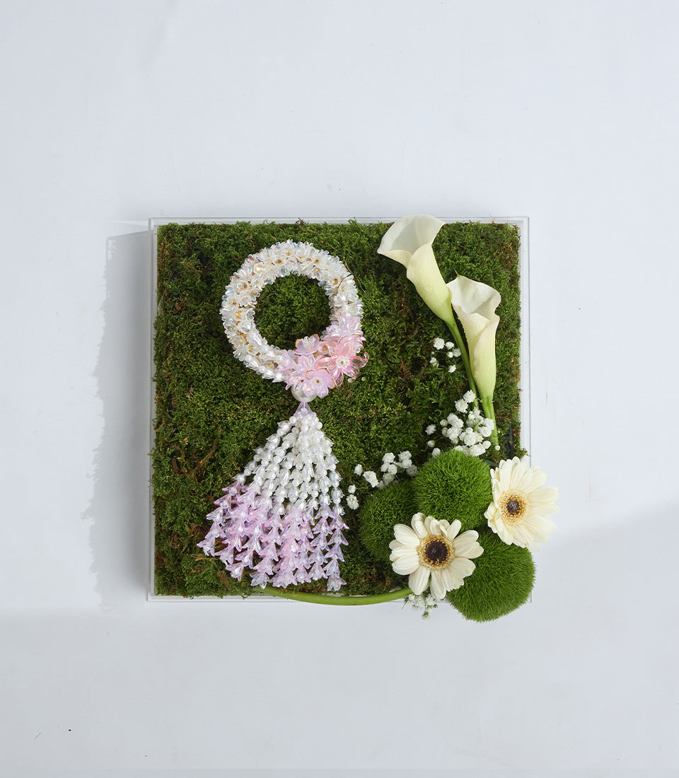 Preserved moss floral art with white and pink flower wreath, cascading lilac blooms, white baby's breath, cream calla lilies, and white gerbera daisies on green moss background.
