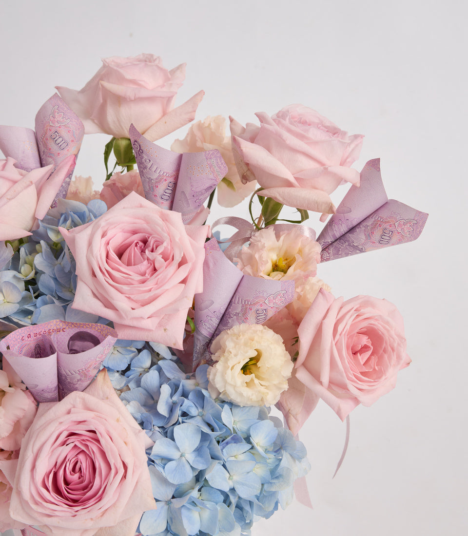Elegant floral bouquet with pink roses, cream carnations, blue hydrangeas, and decorative purple ribbon bows against a white background.