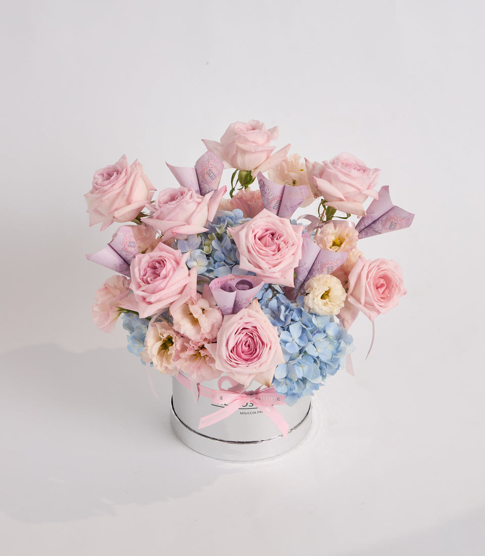 Luxurious floral arrangement in white cylindrical box with pink roses, blue hydrangeas, peach peonies, and purple foliage accents with pink ribbon.
