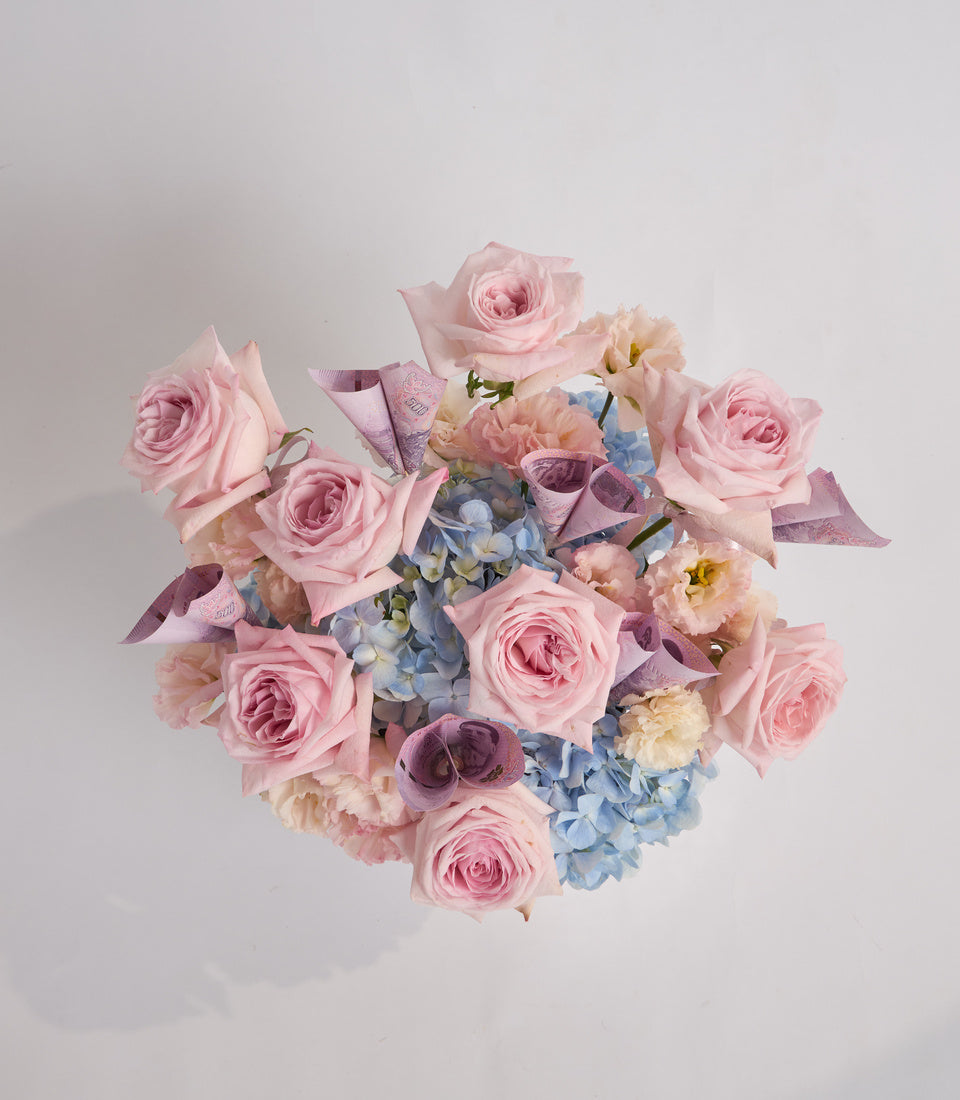 Luxurious bouquet of pink roses, cream peonies, blue hydrangeas, and purple eucalyptus with ribbon accents on white background.