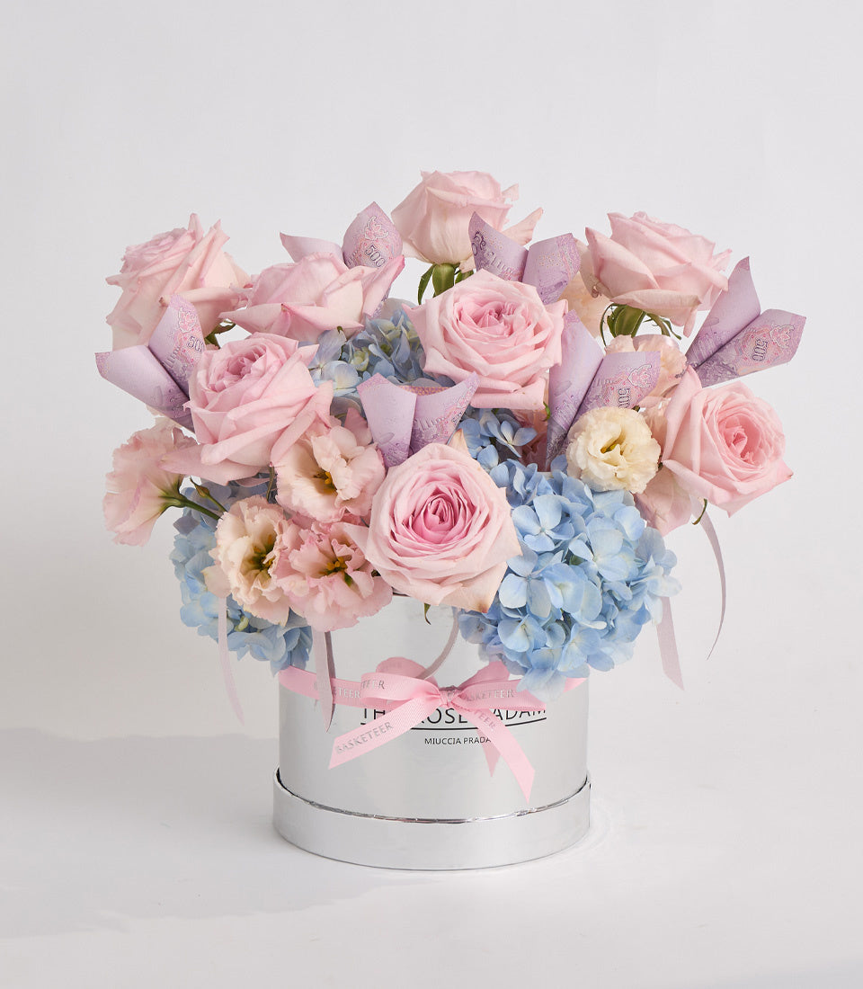 Luxury floral arrangement in white cylindrical box with pink roses, blue hydrangeas, cream carnations, and purple ribbon bow on white background.