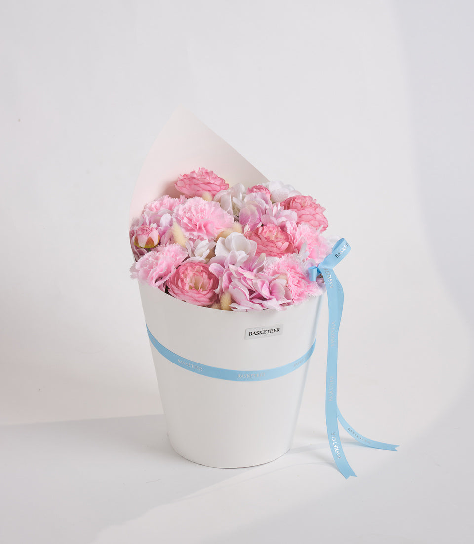 Pink and white peonies in a white cylindrical box with light blue ribbon, branded "Basketeer," against a white background.