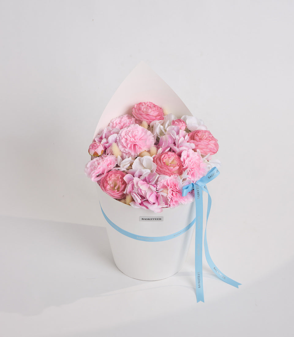 Pink and white peonies in a white cylindrical box with light blue ribbon and "Basketeer" label, elegant floral arrangement.