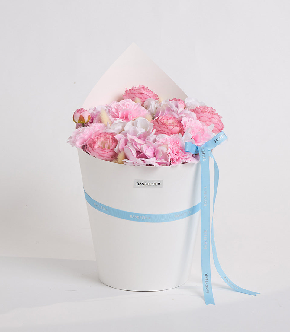 Pink and white peonies in a white cylindrical box with blue "Basketeer" ribbon and tissue paper, displayed against a white background.