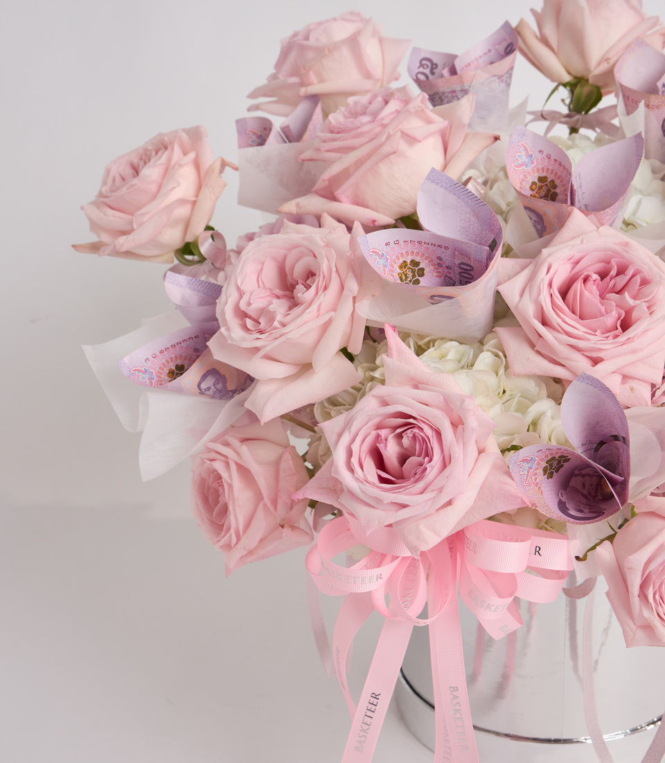 Luxurious floral arrangement with pink roses, white hydrangeas, purple currency accents, and pink ribbons in a white vase.