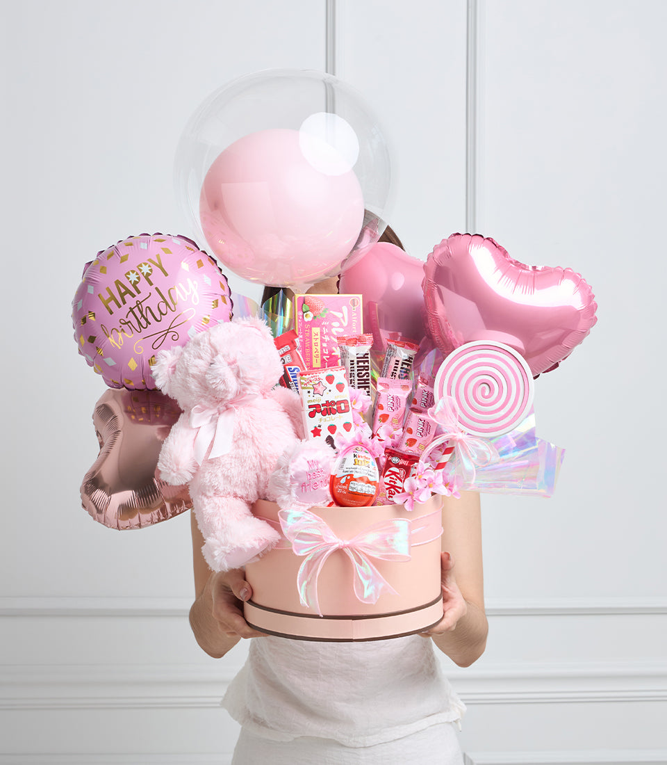 Pink birthday gift box arrangement with balloons, plush teddy bear, candy, and decorative elements held by person in white shirt.