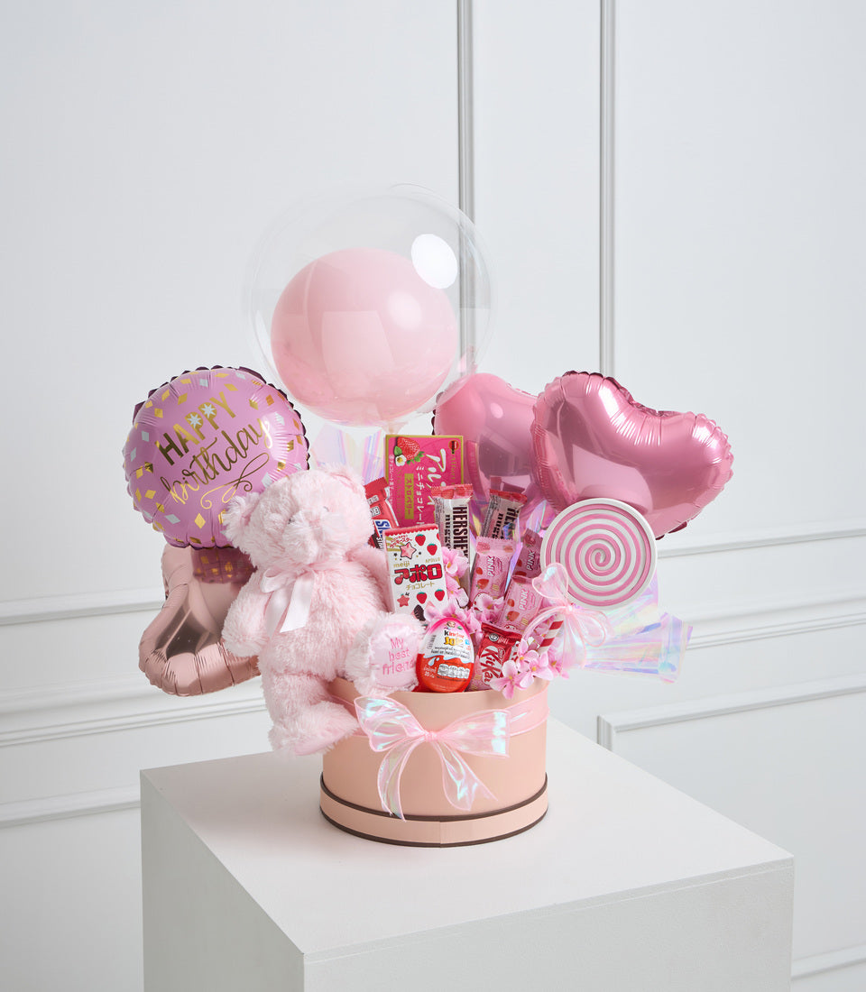 Pink birthday gift arrangement with balloons, plush teddy bear, candy, lollipop decoration, and pink ribbon bow in cylindrical box.