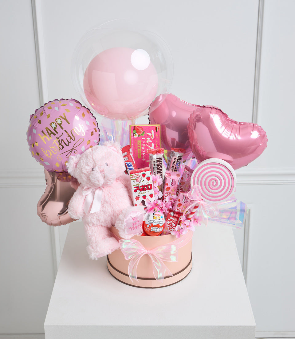 Pink birthday gift basket with balloons, teddy bear, candies, and chocolates in a beige box with iridescent bow.