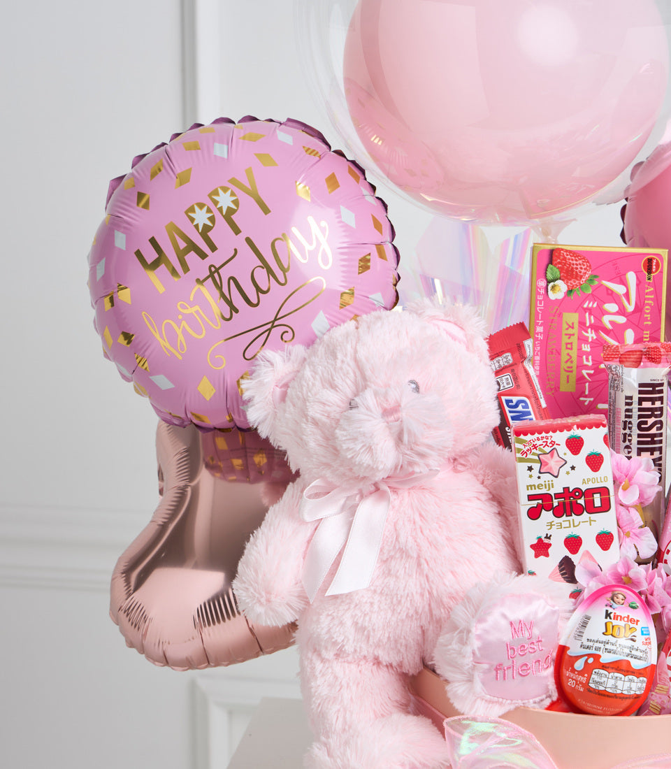 Pink plush teddy bear holding a "Happy Birthday" balloon, surrounded by candy, chocolates, and gift items in pink and red tones.