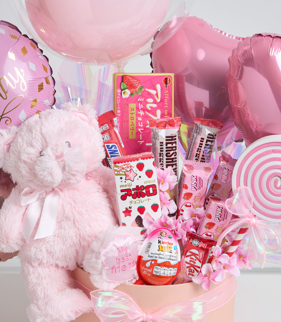Pink themed gift basket with plush teddy bear, balloons, candy assortment, and decorative flowers against white background.