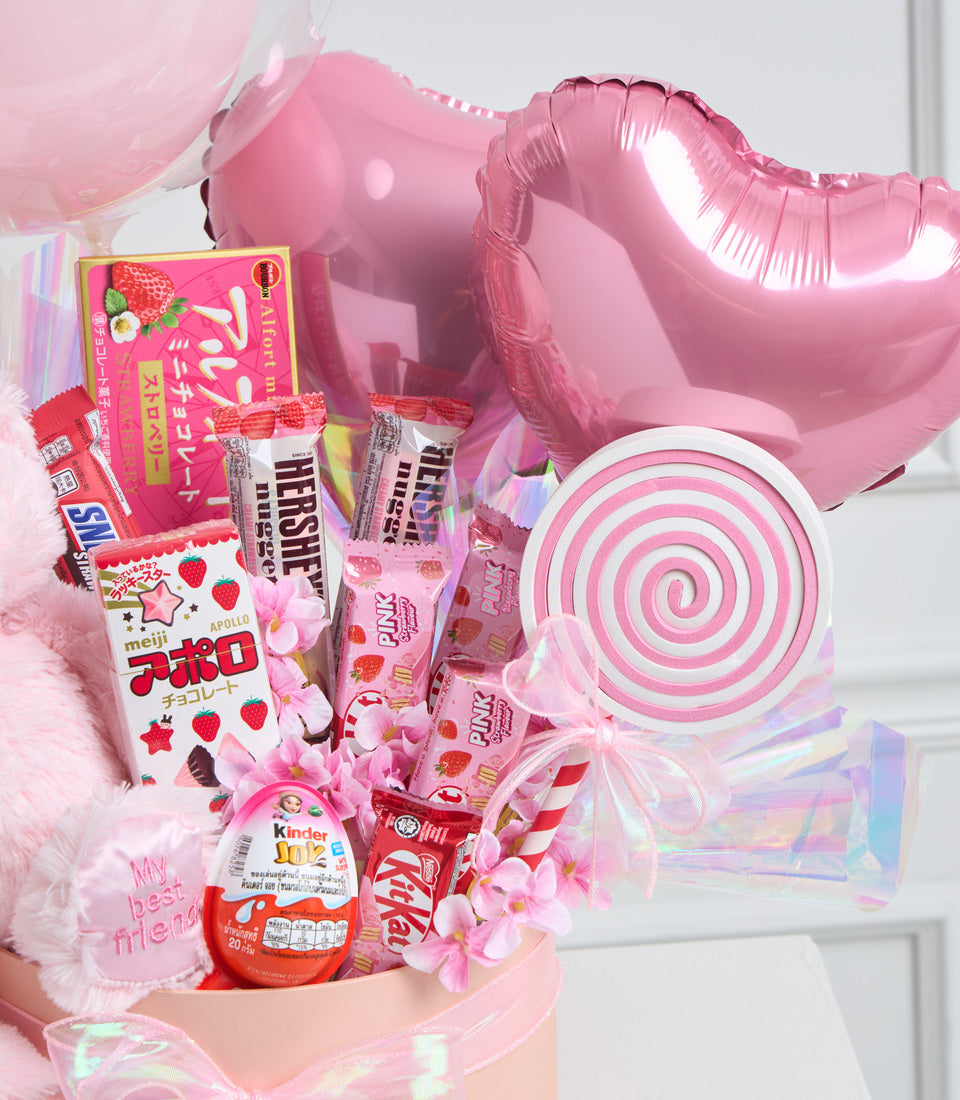 Pink candy gift box arrangement with balloons, lollipop, Hershey's chocolates, Kinder Joyegg, and decorative flowers on white background.