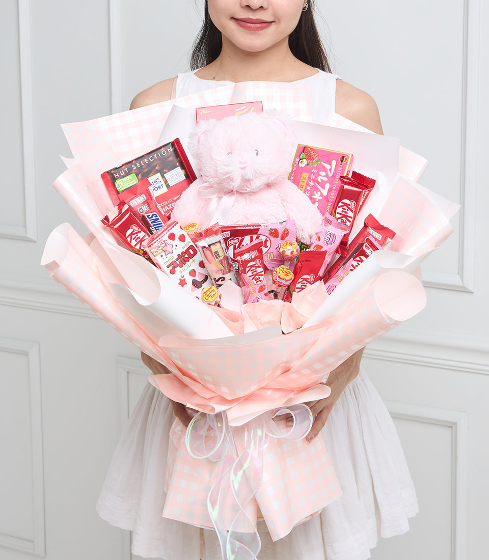 Pink gift bouquet with plush teddy bear, assorted red and pink chocolates, candies, and snacks arranged in layered tissue paper wrapping.