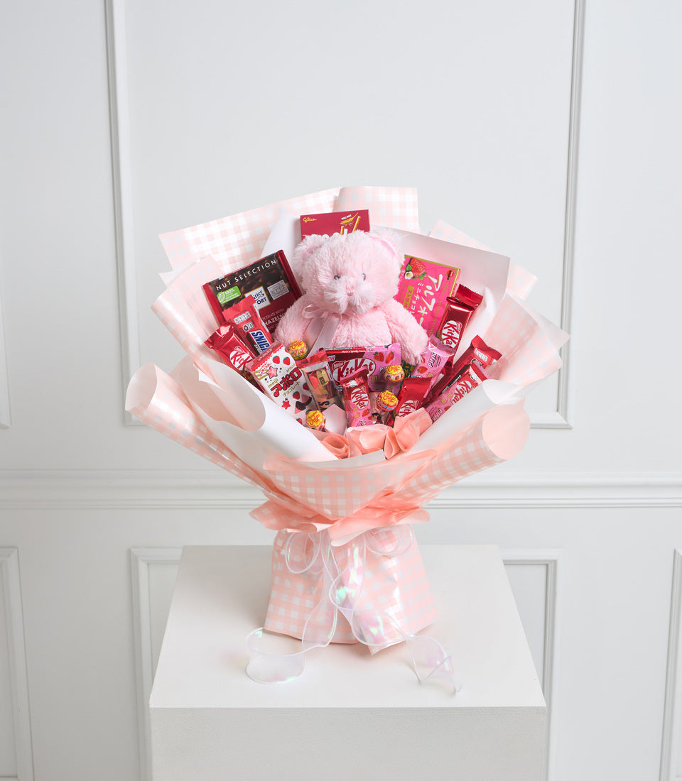 Pink and white checkered bouquet with plush teddy bear, assorted chocolates, candies, and red gift boxes on white pedestal.