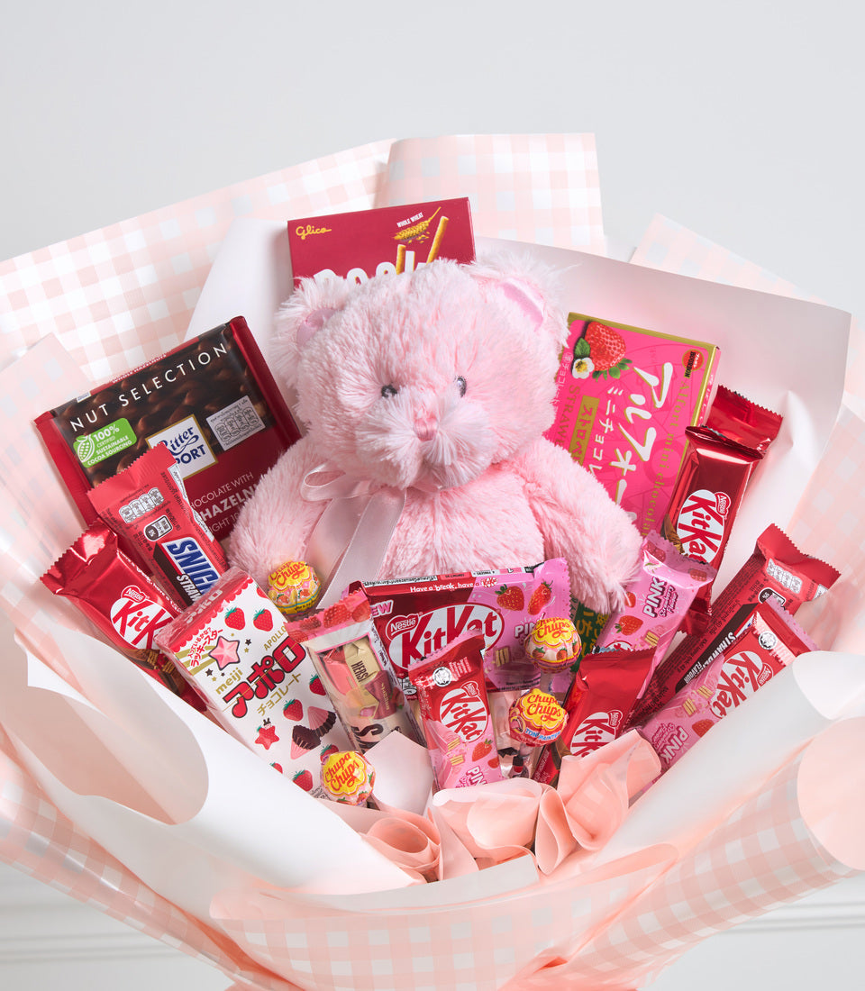 Pink fluffy teddy bear surrounded by assorted red and pink confectionery, chocolates, and sweets arranged in white tissue paper wrapping with pink ribbons.