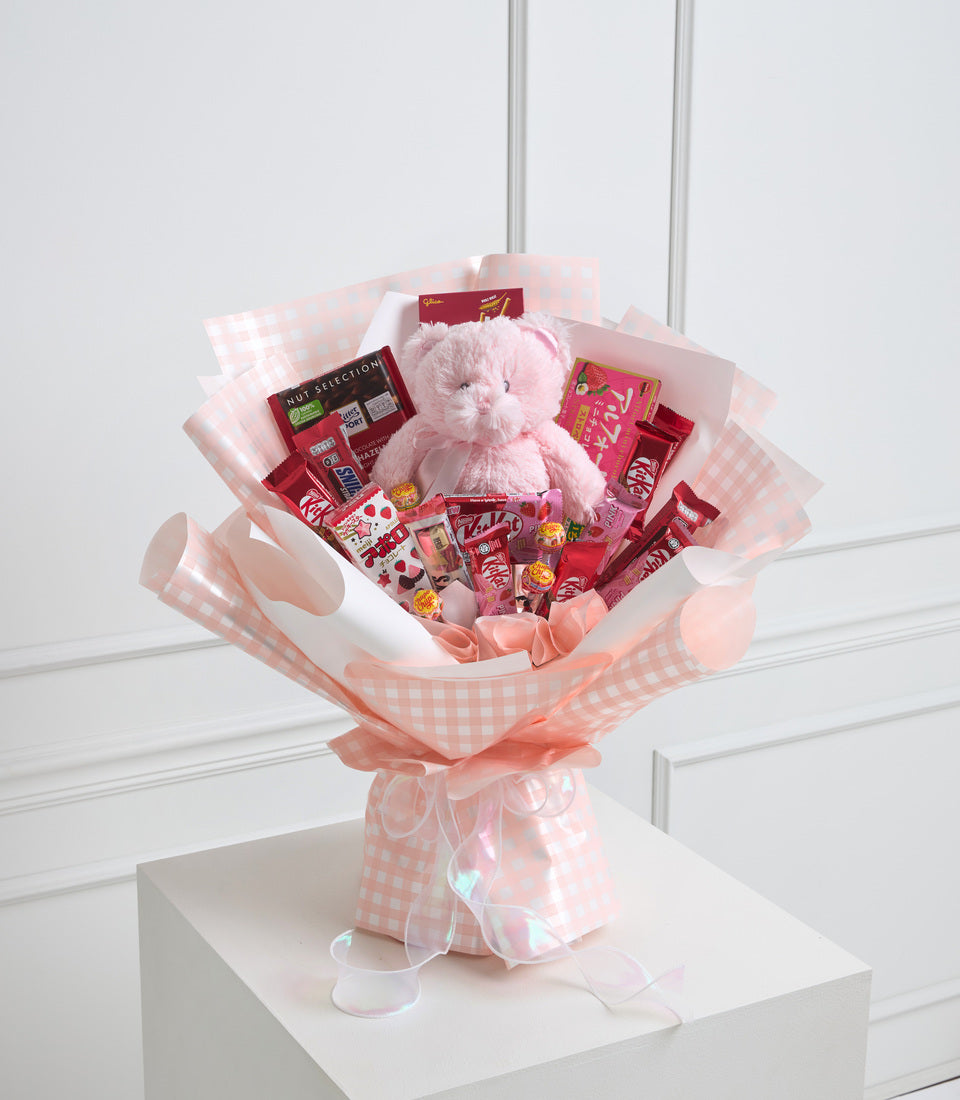 Pink checkered gift bouquet with plush teddy bear, chocolates, candies, and assorted treats wrapped in tissue paper on white pedestal.