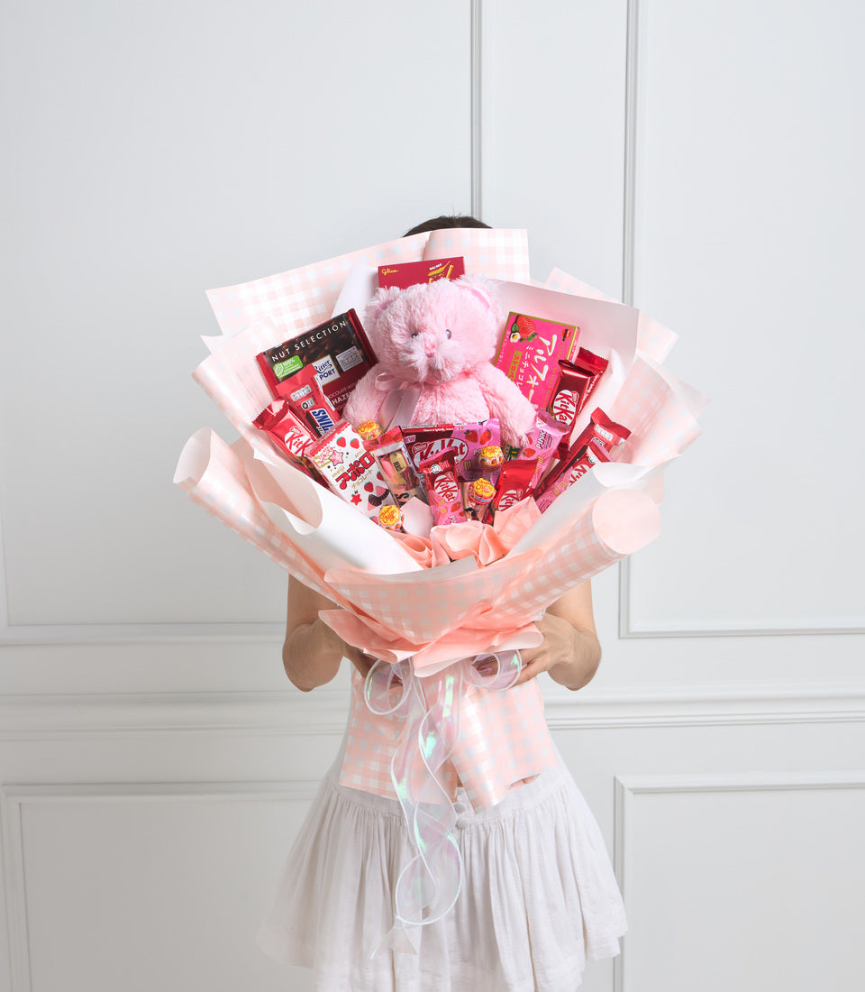 Pink and red candy bouquet with plush teddy bear, chocolate bars, and sweets wrapped in layered pink checkered and white tissue paper.