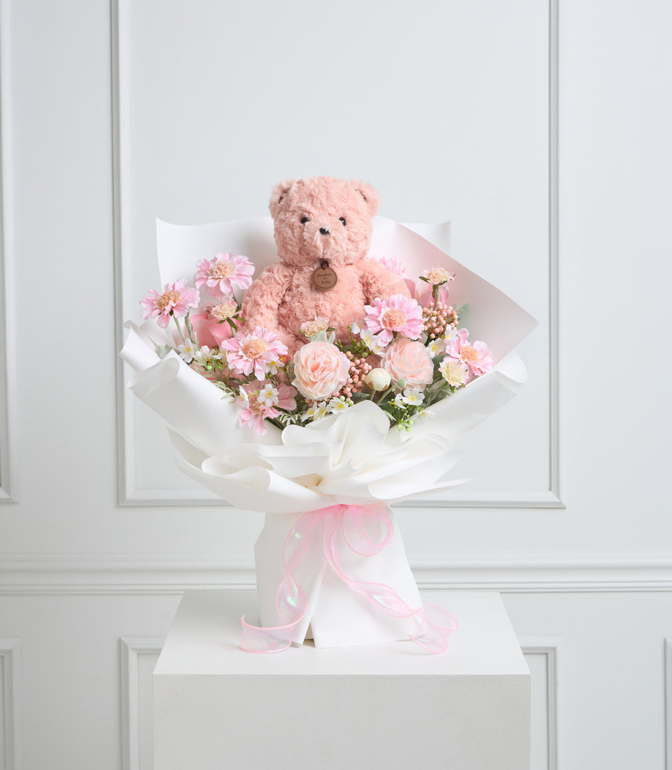 Pink teddy bear sitting in a bouquet of pink and white flowers with white wrapping and pink ribbon bow, displayed on white pedestal.