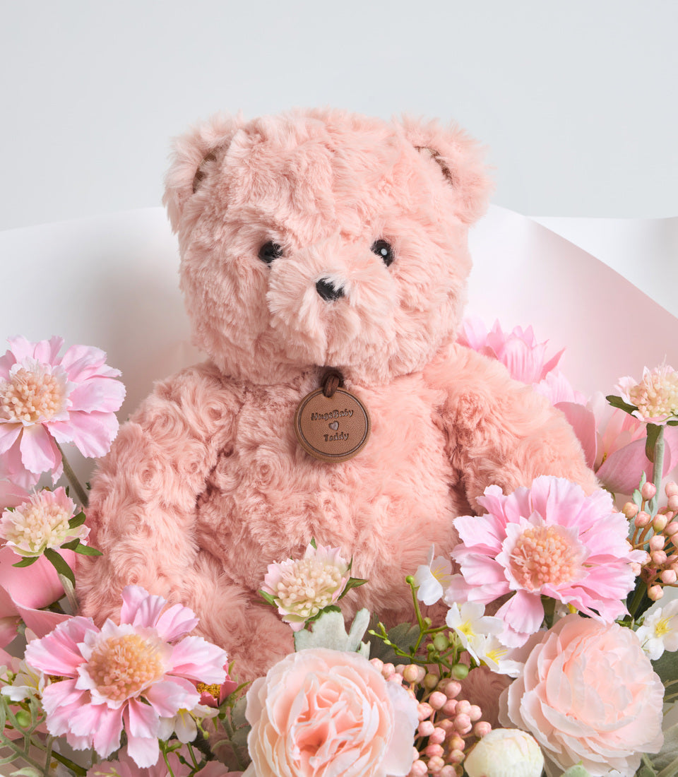 Pink fluffy teddy bear with engraved heart pendant surrounded by pink flowers including gerberas and roses.