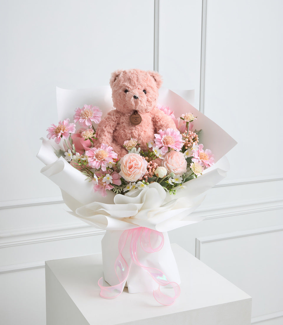 Pink teddy bear surrounded by pink and peach flowers with white wrapping and pink ribbon bow, elegant gift arrangement.