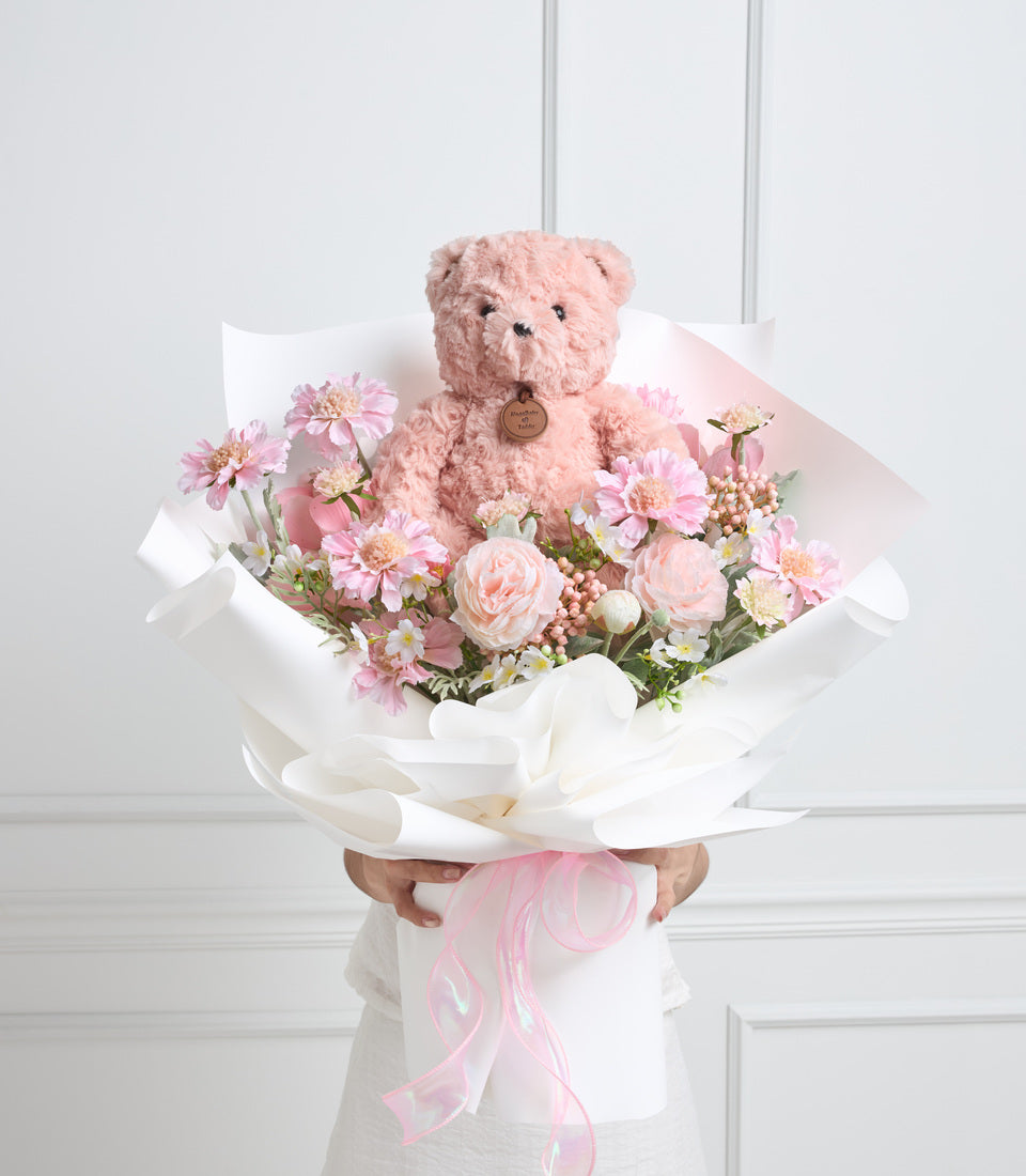 Pink teddy bear surrounded by pink and white flowers including gerberas and roses, wrapped in white paper with pink ribbon.