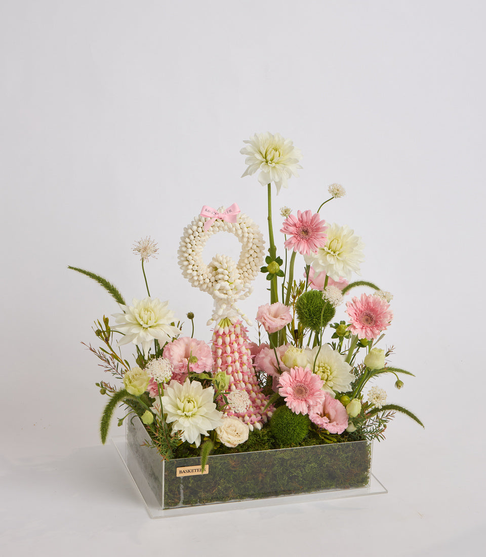 Elegant floral arrangement featuring cream and pink gerberas, dahlias, and carnations in a gray rectangular planter with moss base and clear acrylic stand.
