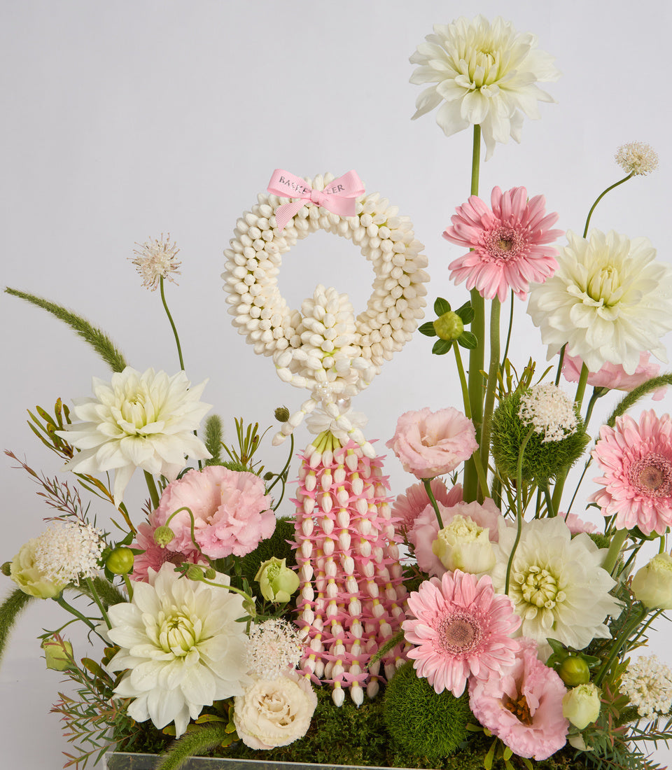 Elegant floral arrangement with white dahlias, pink gerberas, pearl beaded number 9 and bow, lisianthus, and greenery on gray background.