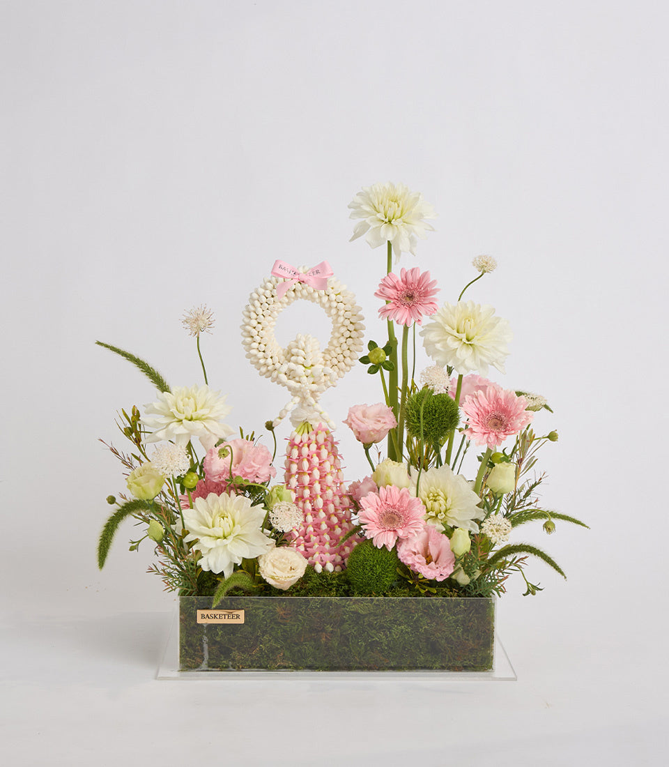 Pink and cream floral arrangement in a textured grey-green rectangular container with moss, featuring gerberas, dahlias, and a pearl beaded wreath accent.