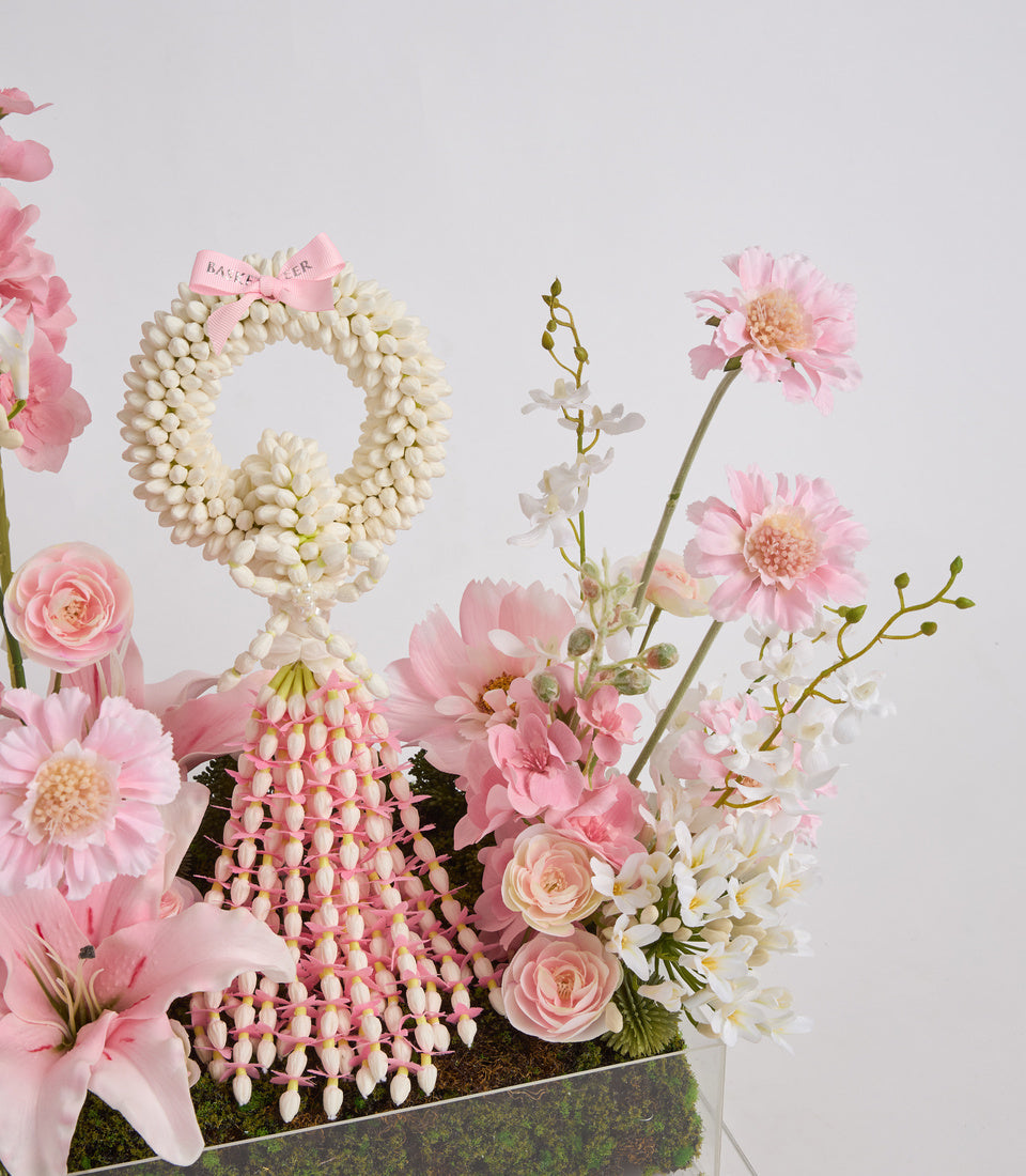 Luxe floral arrangement with pink gerberas, roses, white daisies, pearl wreath with pink ribbon, and green moss in modern vase.