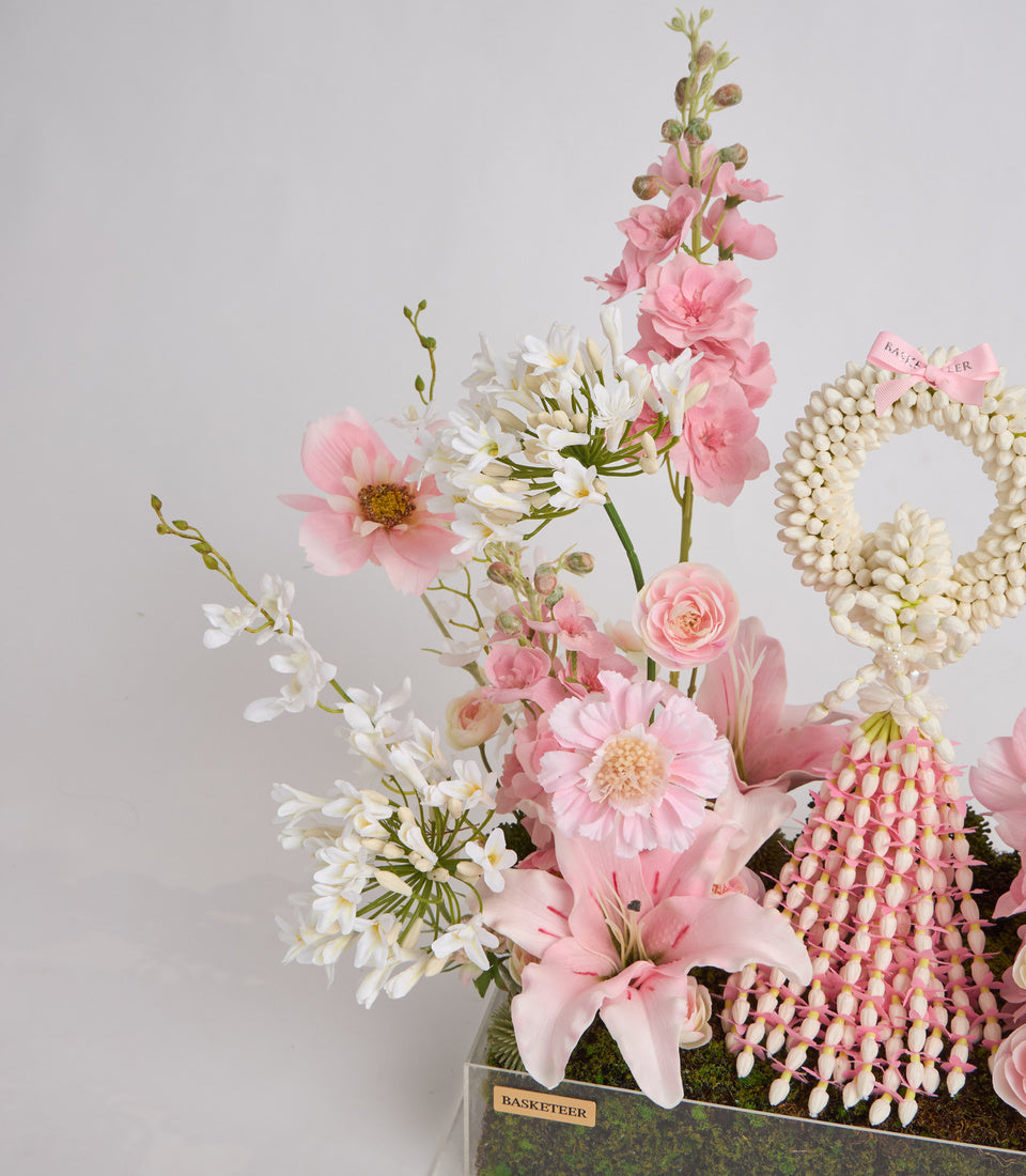 Luxe floral arrangement with pink lilies, roses, white daisies, pearl beads, and decorative elements in a branded container.