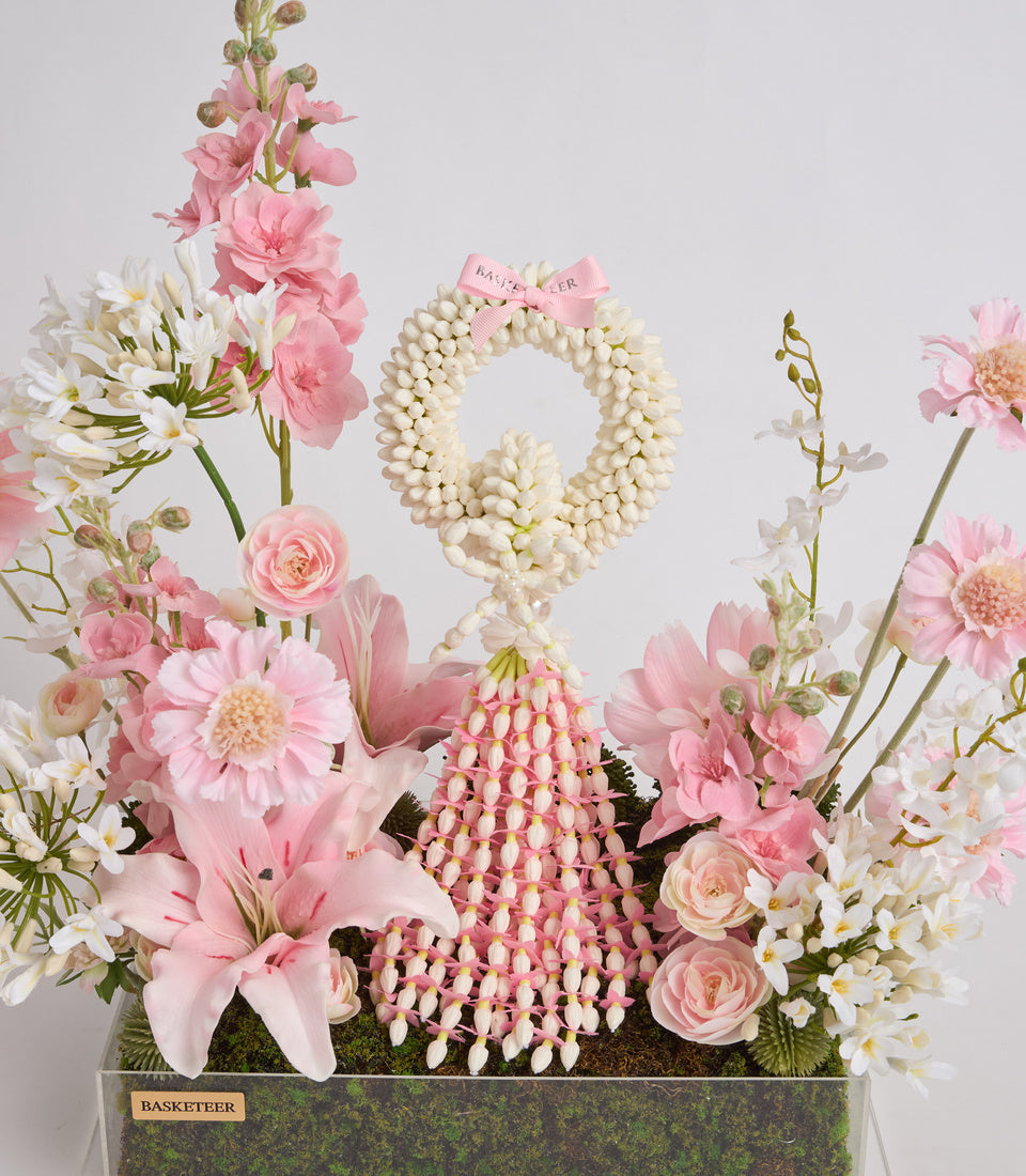 Luxurious floral arrangement in moss-green vase featuring pink roses, white daisies, lilies, pearl beaded wreath, and pink ribbon bow on neutral background.