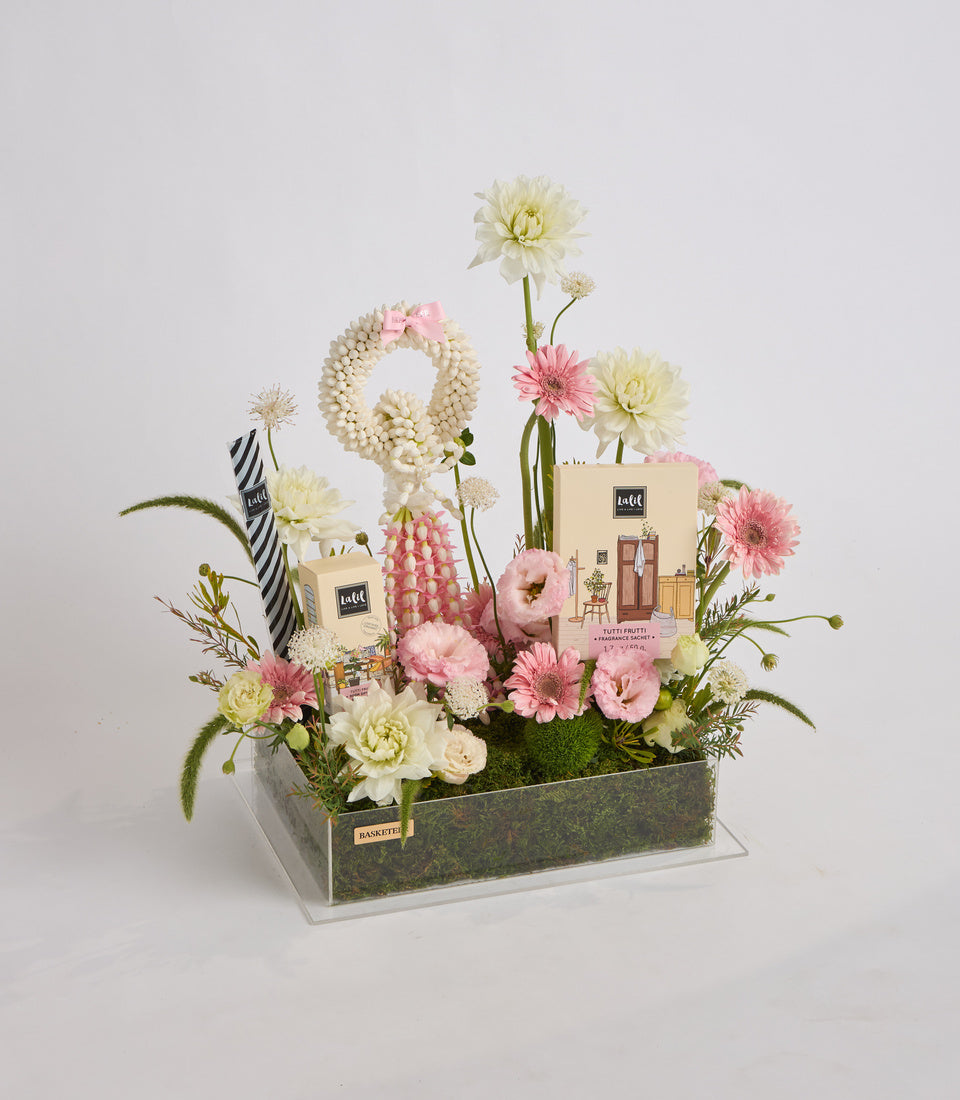 Luxe floral arrangement in rectangular green container with pink and cream flowers, beaded pearl garland, striped ribbon, and Laffi fragrance products displayed among blooms.