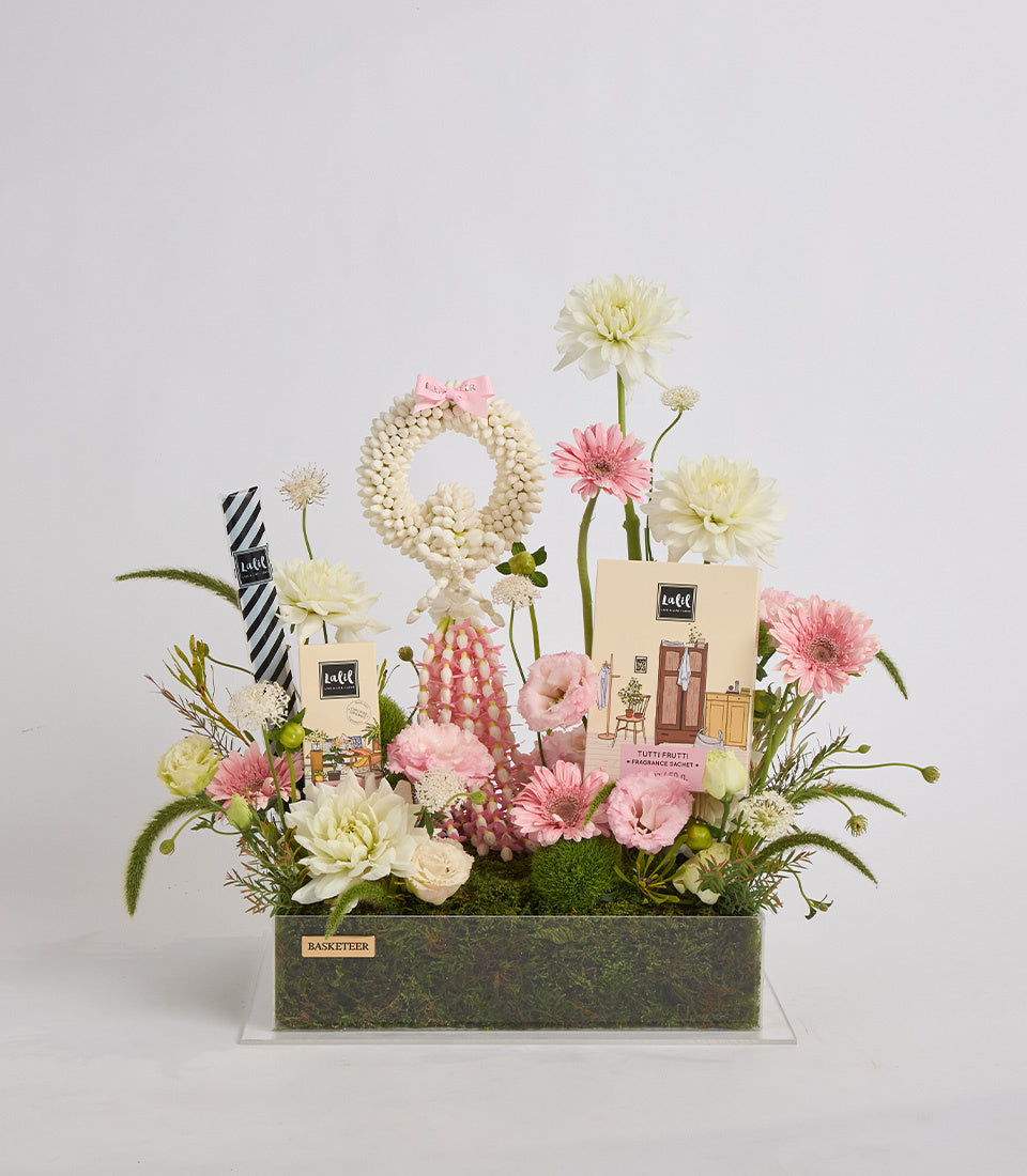 Luxe floral arrangement featuring cream and pink flowers, beaded wreath, fragrance boxes, striped ribbon, and moss in a textured container.