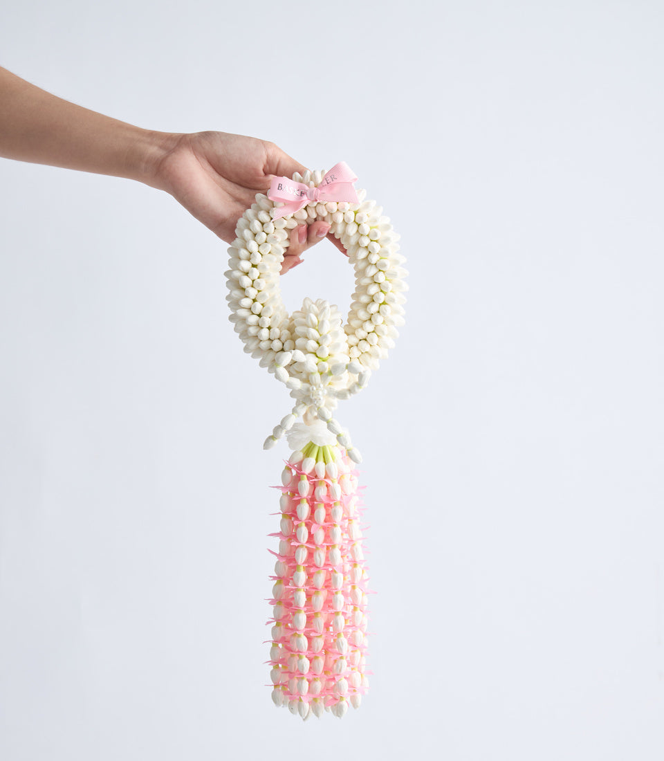 Hand holding a jasmine flower garland lei with cream-colored beads and pink flowers, featuring a pink bow at the top and cascading pink blooms below.
