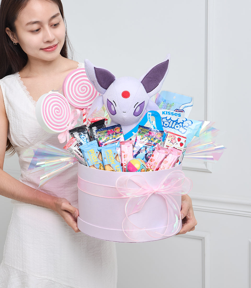 Woman holding pink cylindrical gift box with purple cat plush, lollipops, candies, and iridescent wings against white background.