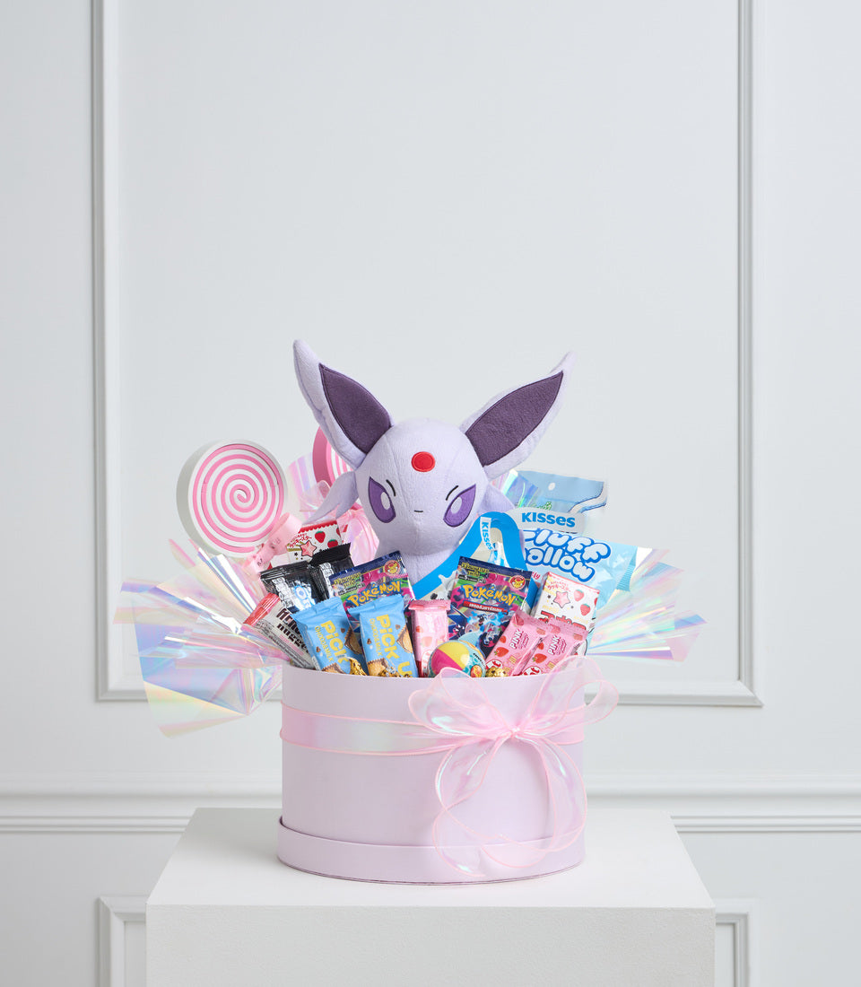 Pink gift box with Pokémon Espeon plush, candies, lollipop, and pastel ribbons on white pedestal against bright background.