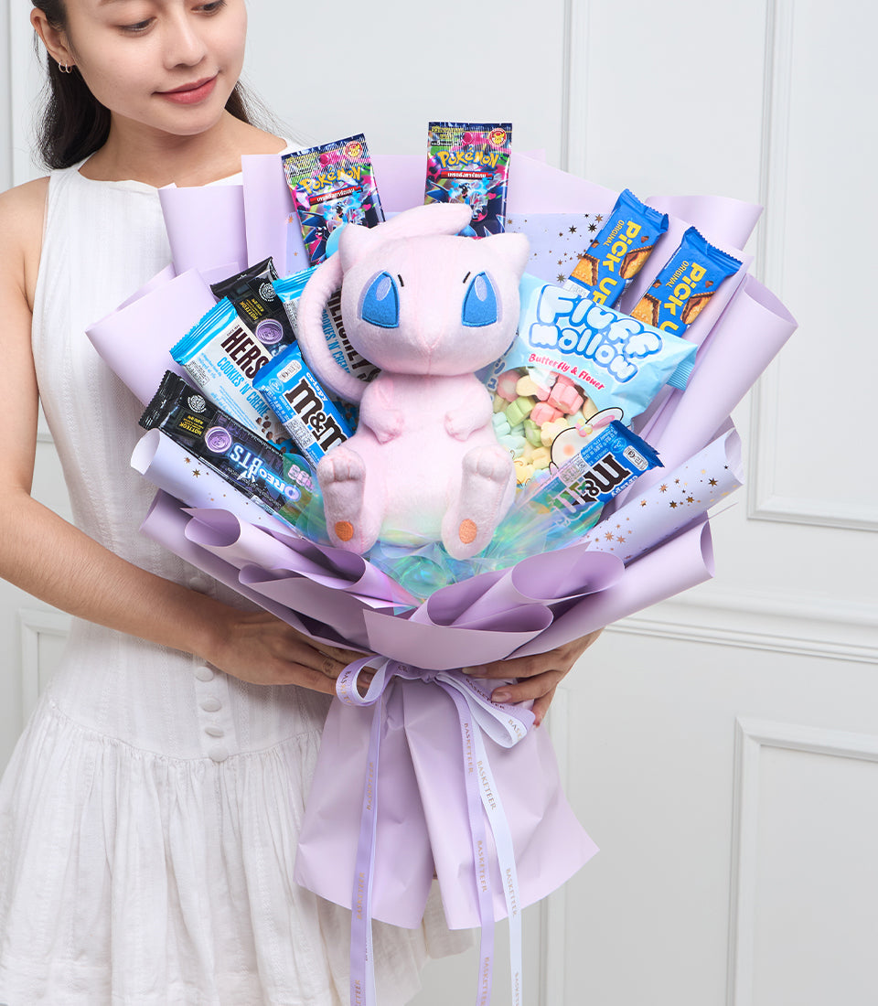 Woman holding purple wrapped bouquet with pink Mew Pokémon plush, candy, trading cards, and snacks arranged among lavender tissue paper.
