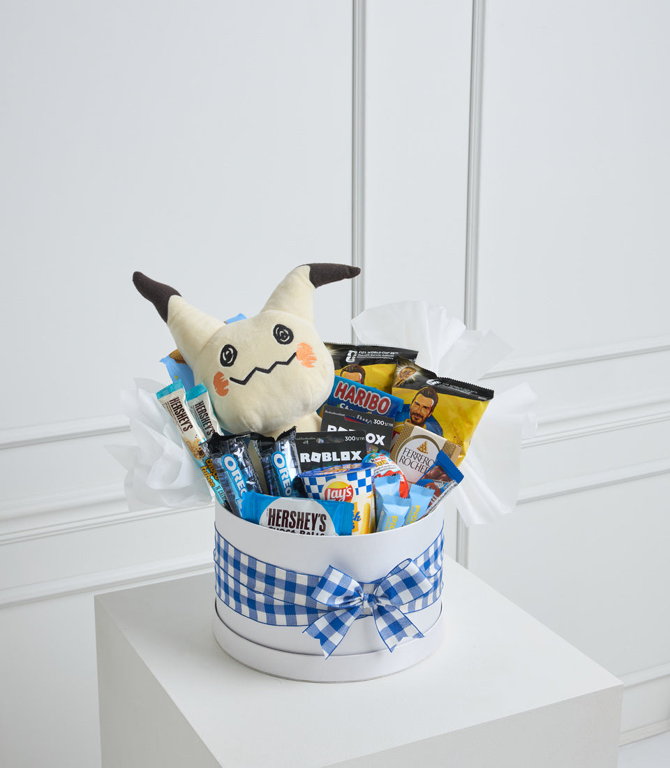 Decorative gift basket with white cylindrical container, blue gingham ribbon bow, filled with snacks and candy, topped with cream and black horned plush Mimikyu character toy.