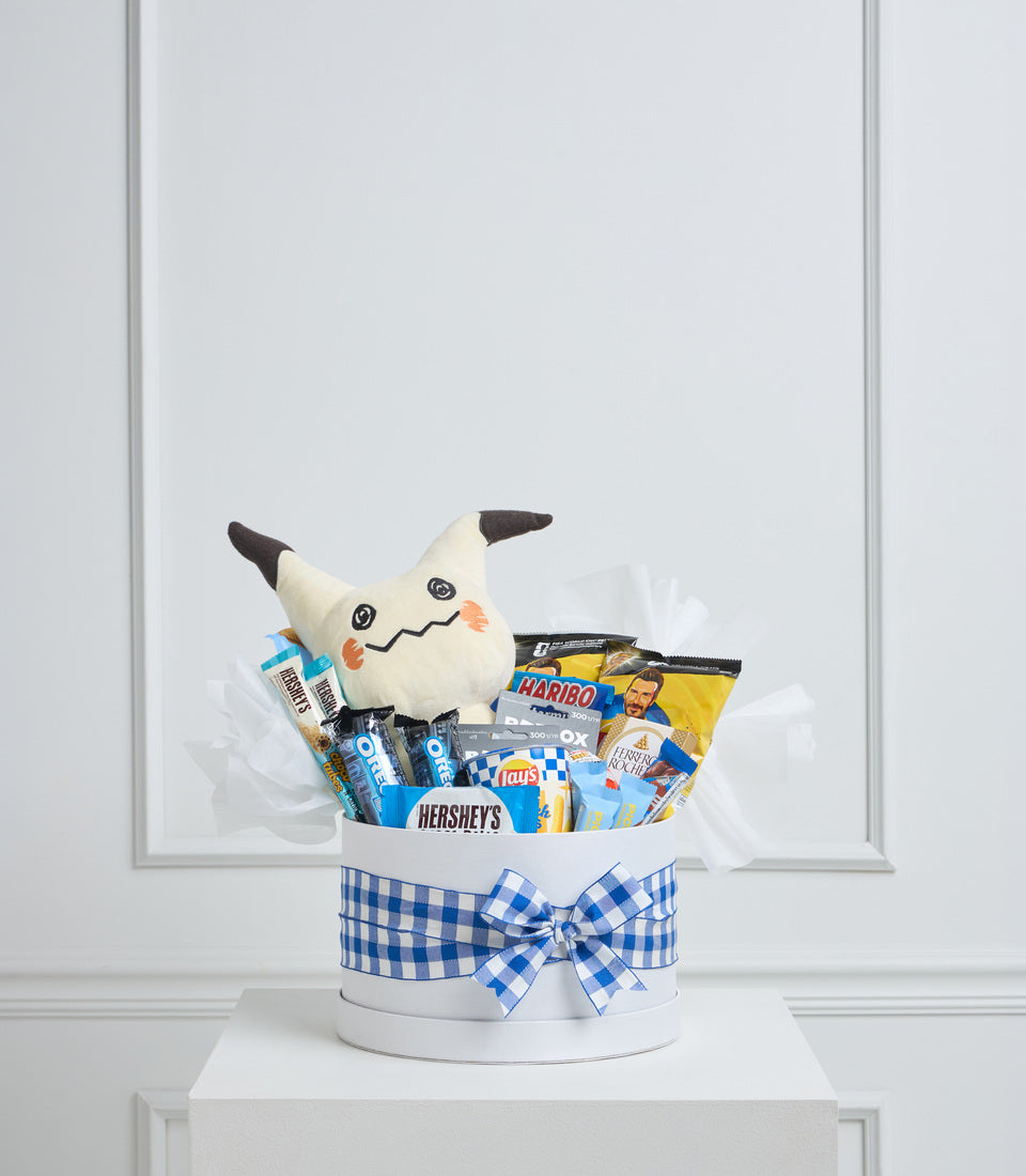 White gift basket with blue gingham bow containing Mimikyu plush toy, chocolates, chips, and assorted snacks on white pedestal.