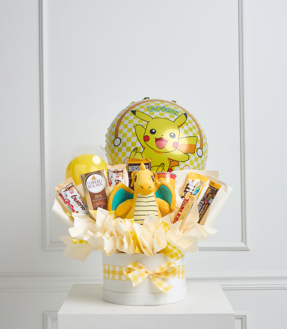 Yellow and white Pokémon gift basket with Pikachu balloon, plush Squirtle, chocolates, beauty products, and gingham ribbon bow on white pedestal.