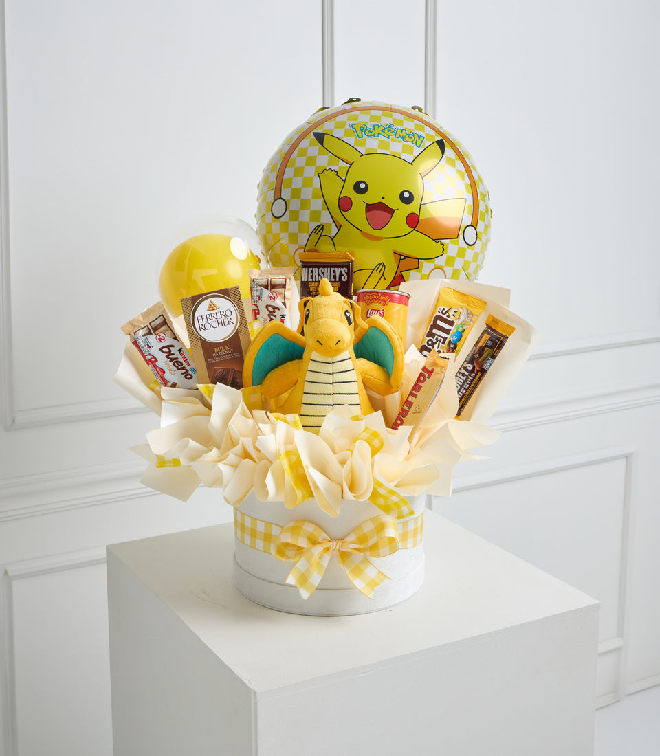 Pokémon-themed gift basket featuring yellow and white colors with Pikachu balloon, plush toy, snacks, chocolates, and yellow checkered bow on white pedestal.