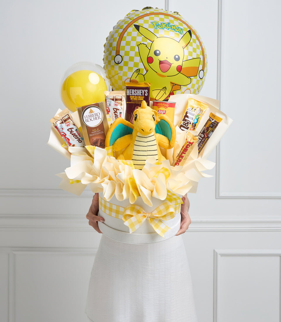 Yellow and white Pokémon gift basket featuring Pikachu balloon, plush Dragonite, snacks, chocolate, and candy arranged with cream tissue paper and yellow gingham bow.