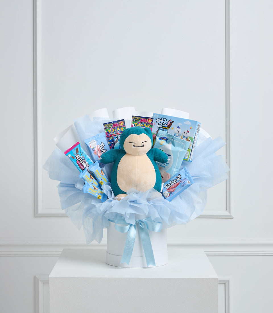 Blue and white gift bouquet featuring a teal Snorlax plush centerpiece surrounded by Pokemon trading cards, candy, and toys arranged in tulle wrapping.