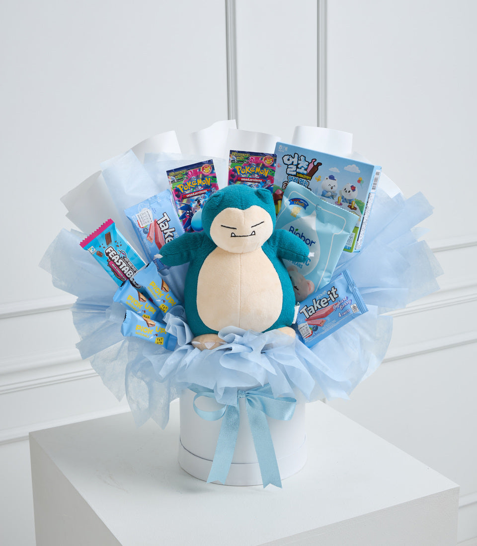 Pokémon Snorlax plush centerpiece surrounded by trading cards, candy, and toys in light blue tulle arrangement with blue ribbon bow.