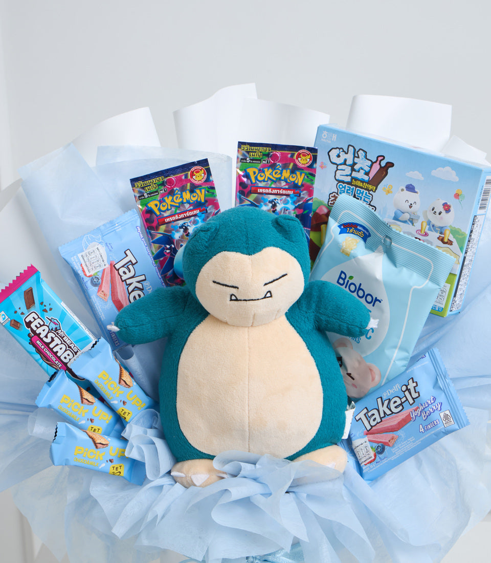 Blue Snorlax plush surrounded by Pokemon magazines and Thai snacks in clear cellophane with light blue ribbon bow.