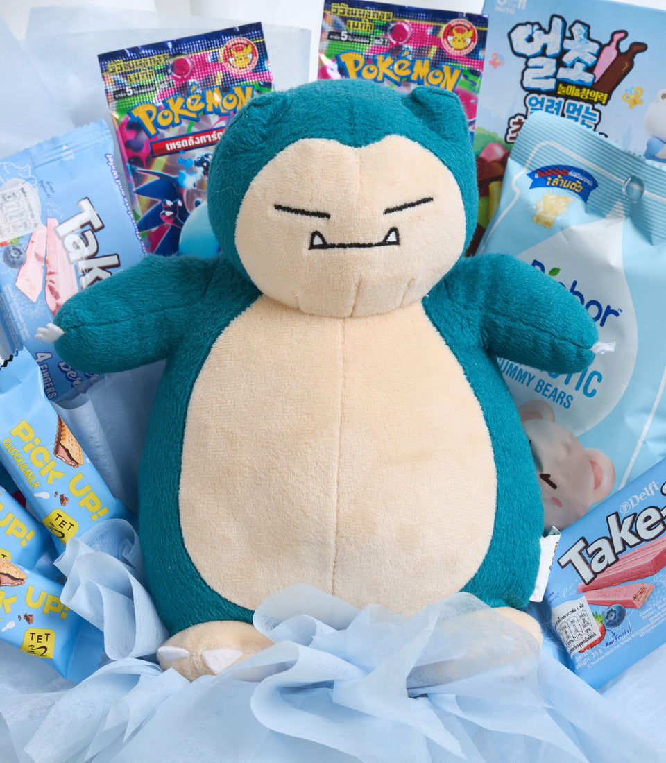 Snorlax plush toy surrounded by Pokemon merchandise and snacks on white tulle, featuring teal and cream colors.