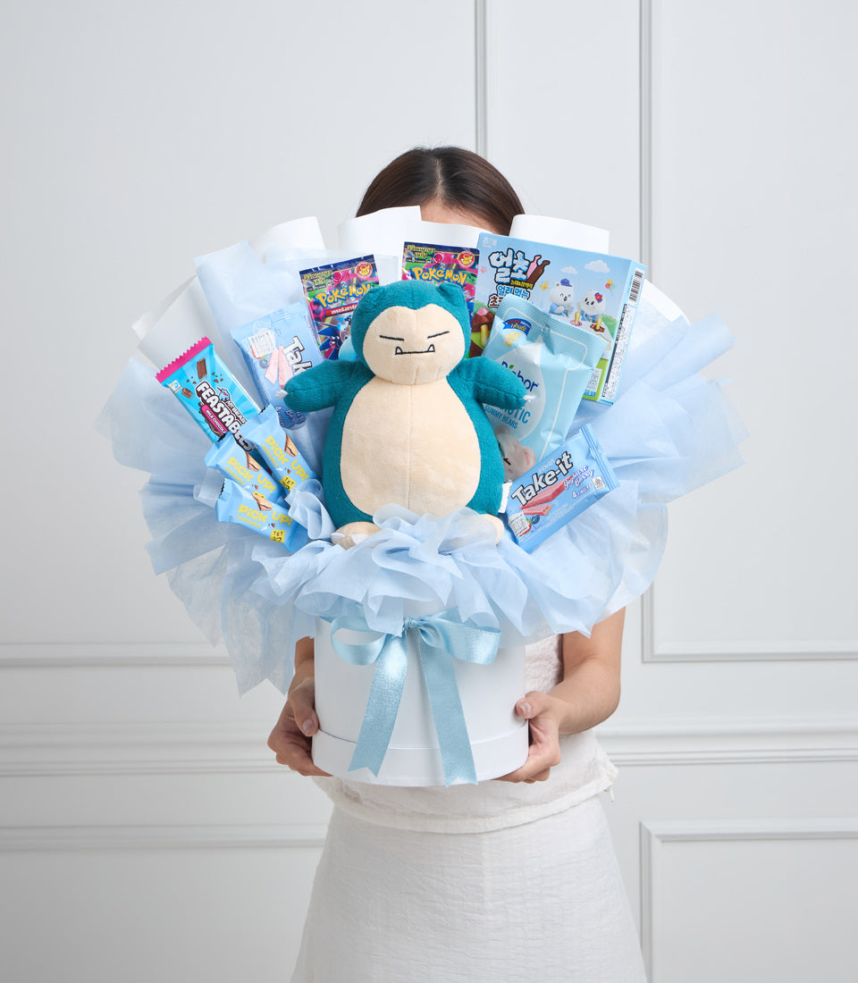 Woman holding a large blue tulle bouquet arrangement with a Snorlax plush center, surrounded by Pokemon-themed candy and toys, tied with a light blue ribbon.