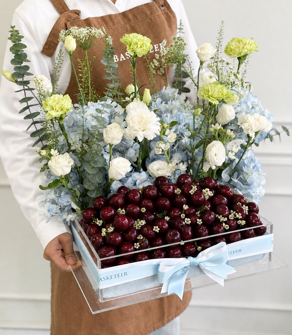 Florist holding luxury gift box with fresh cherries, white and yellow carnations, blue hydrangeas, eucalyptus, and baby's breath with light blue ribbon.
