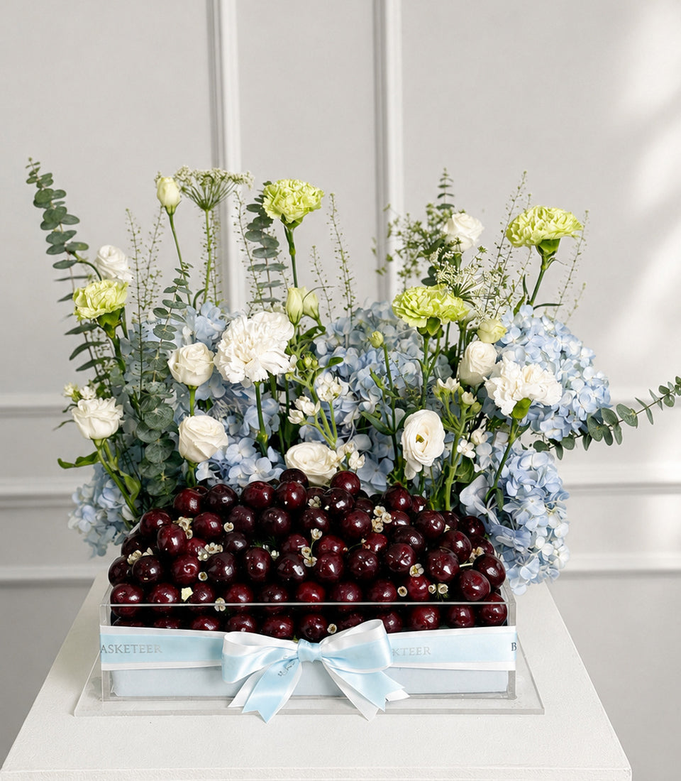 Luxury gift box with fresh dark cherries, white and pale yellow carnations, blue hydrangeas, white roses, and eucalyptus, tied with light blue ribbon bow.