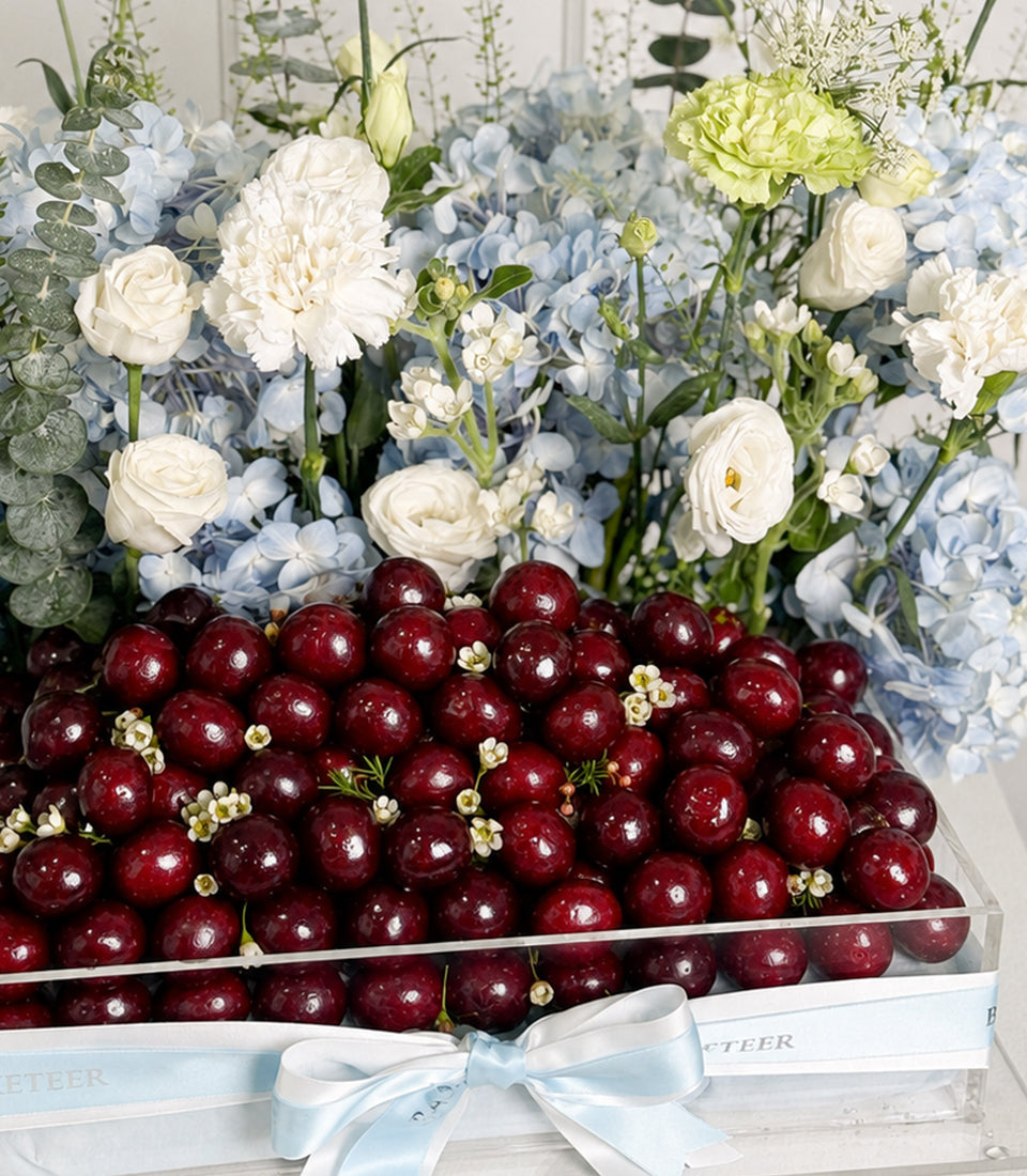 Luxury gift box with dark red cherries, white roses, pale yellow carnations, blue hydrangeas, and eucalyptus, tied with light blue ribbon.